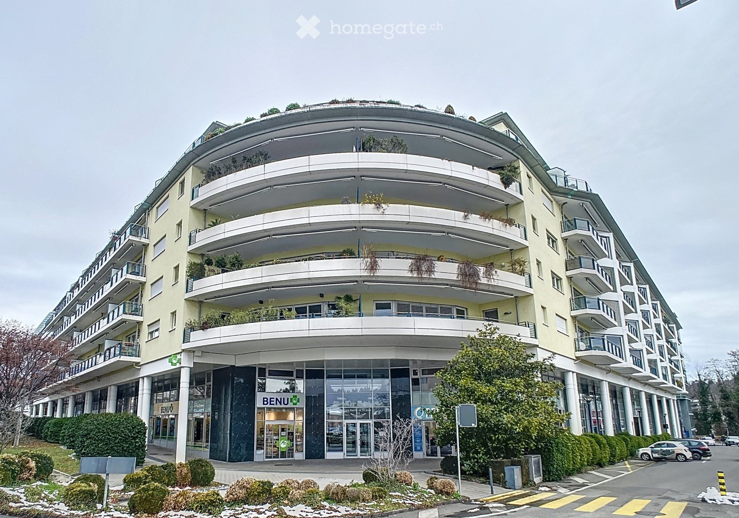 multi-story building, yellow exterior, glass windows, multiple balconies, ground floor with shops, green plants on balconies