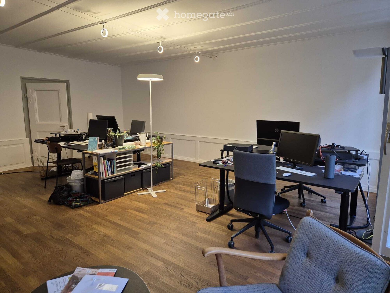 Office room with wooden floor, white walls, ceiling lights, desks, chairs, monitors, keyboards, and plants.