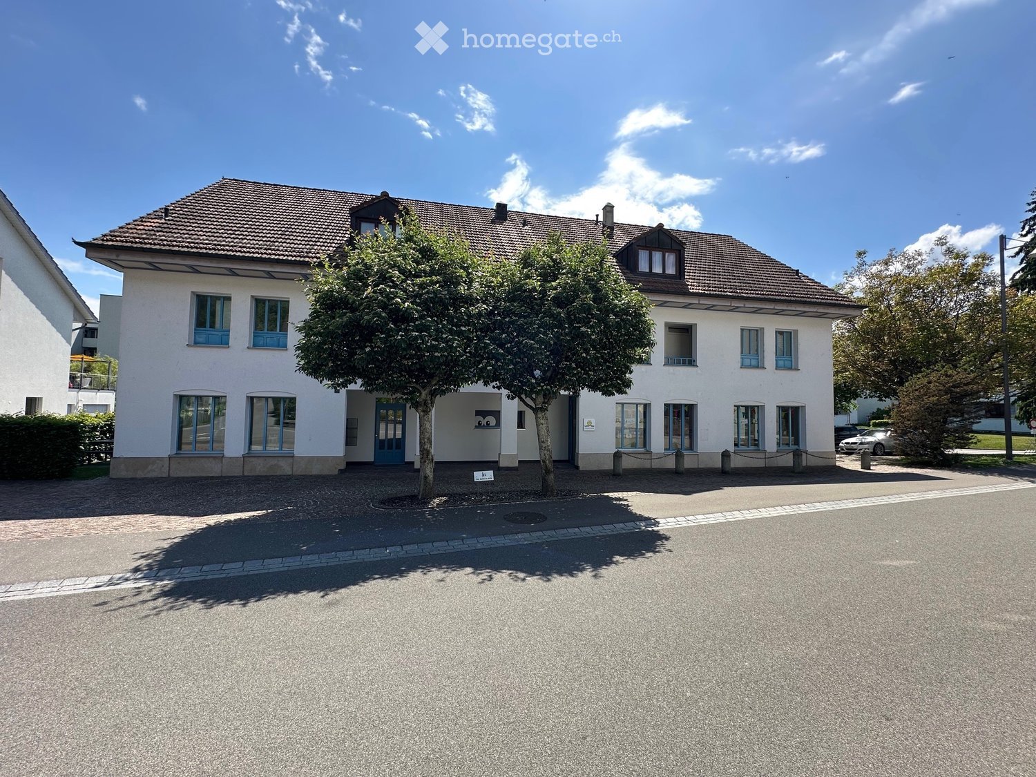 white building, brown roof, multiple windows, two trees in front, paved road, parked car