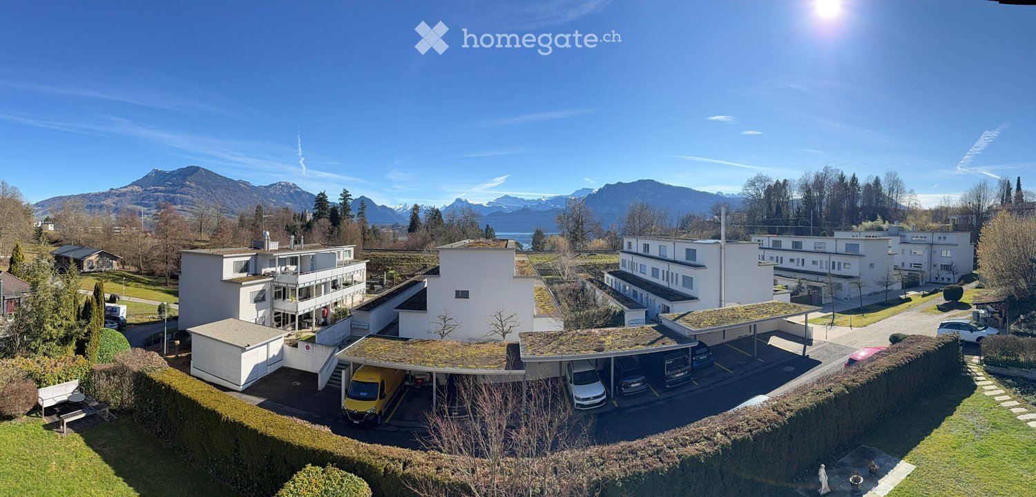 Modern apartment building with covered parking, large windows, green rooftops, several vehicles parked, surrounded by trees and shrubs, mountains in the background.