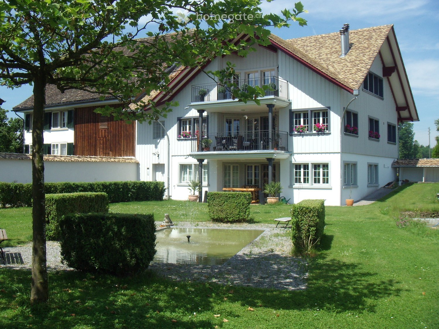 2 story house, white exterior, brown roof, balcony, garden, fountain, hedges, trees