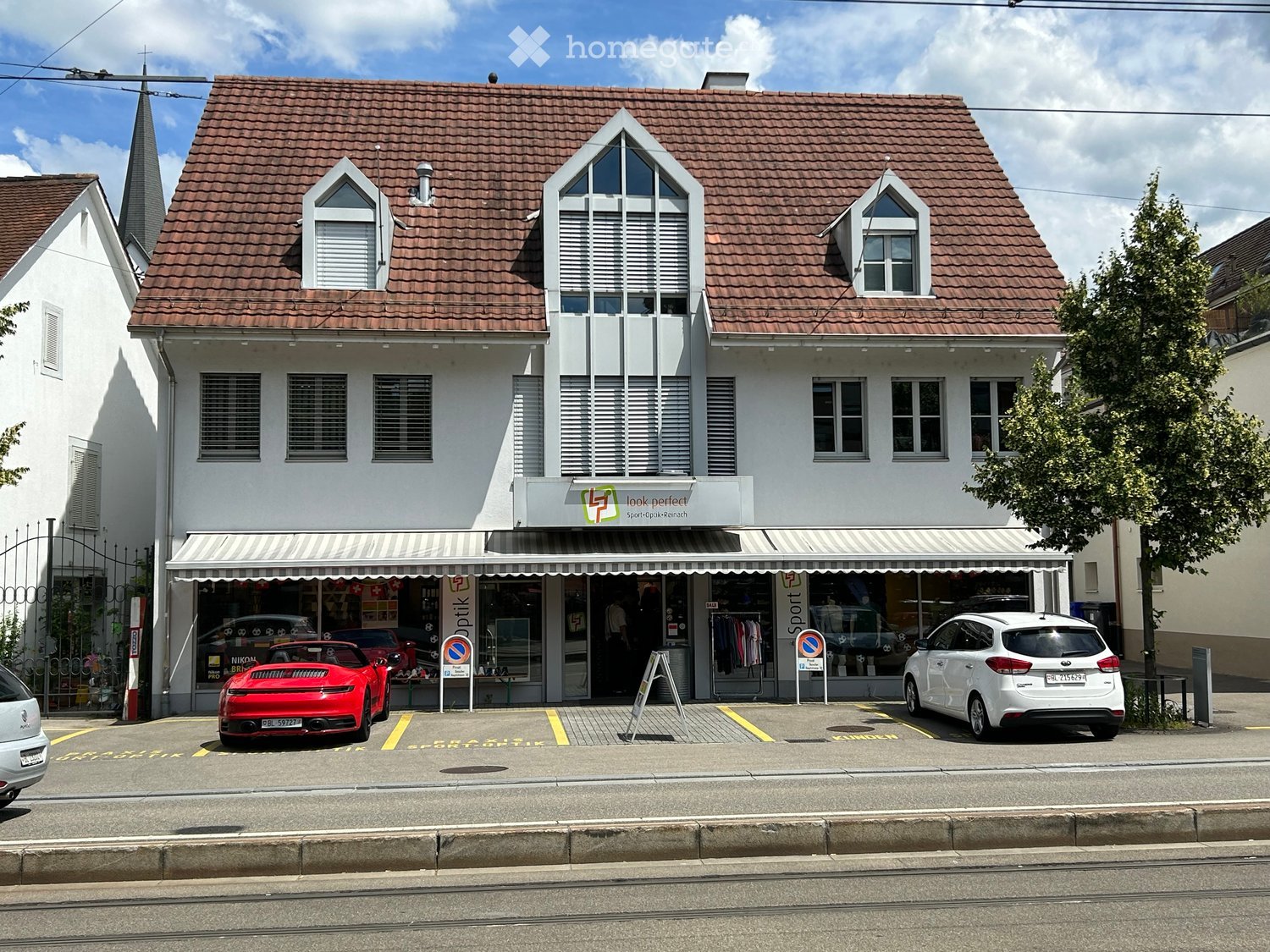2 story house, red roof, white walls, awning, 2 cars parked in front, shop on the ground floor