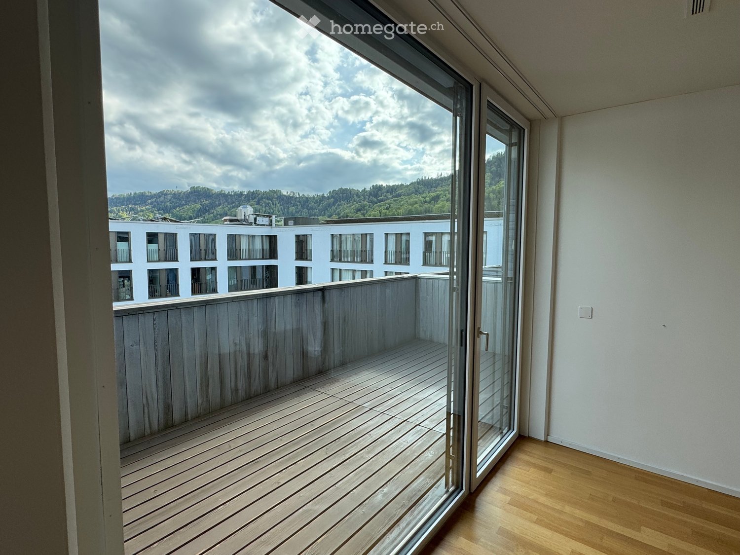 Balcony with wooden floor, sliding glass doors, white walls, view of a building and mountains