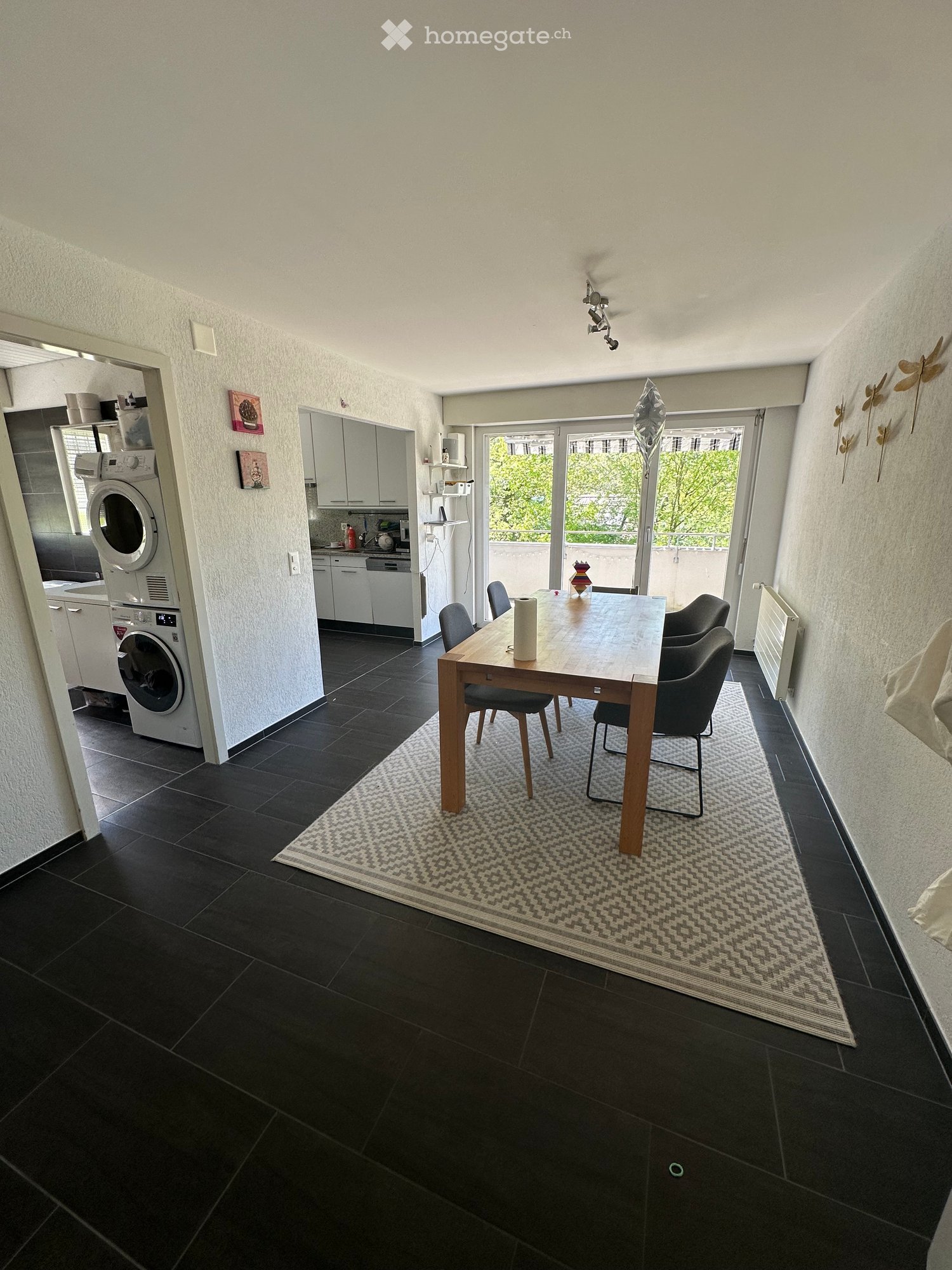 Dining room with wooden table, chairs, rug, and kitchen appliances visible in the background.