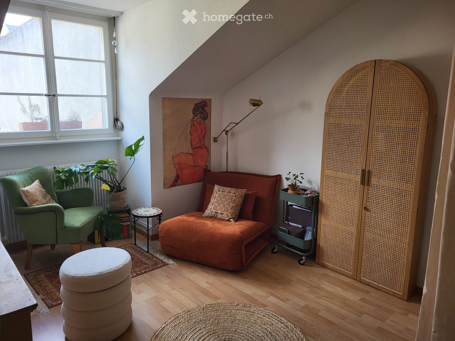 A cozy room with a sofa, a green armchair, a wooden floor, a radiator, a window with a view, a painting, a lamp, a rug, a side table, a stack of books, a potted plant, and a wooden cabinet.