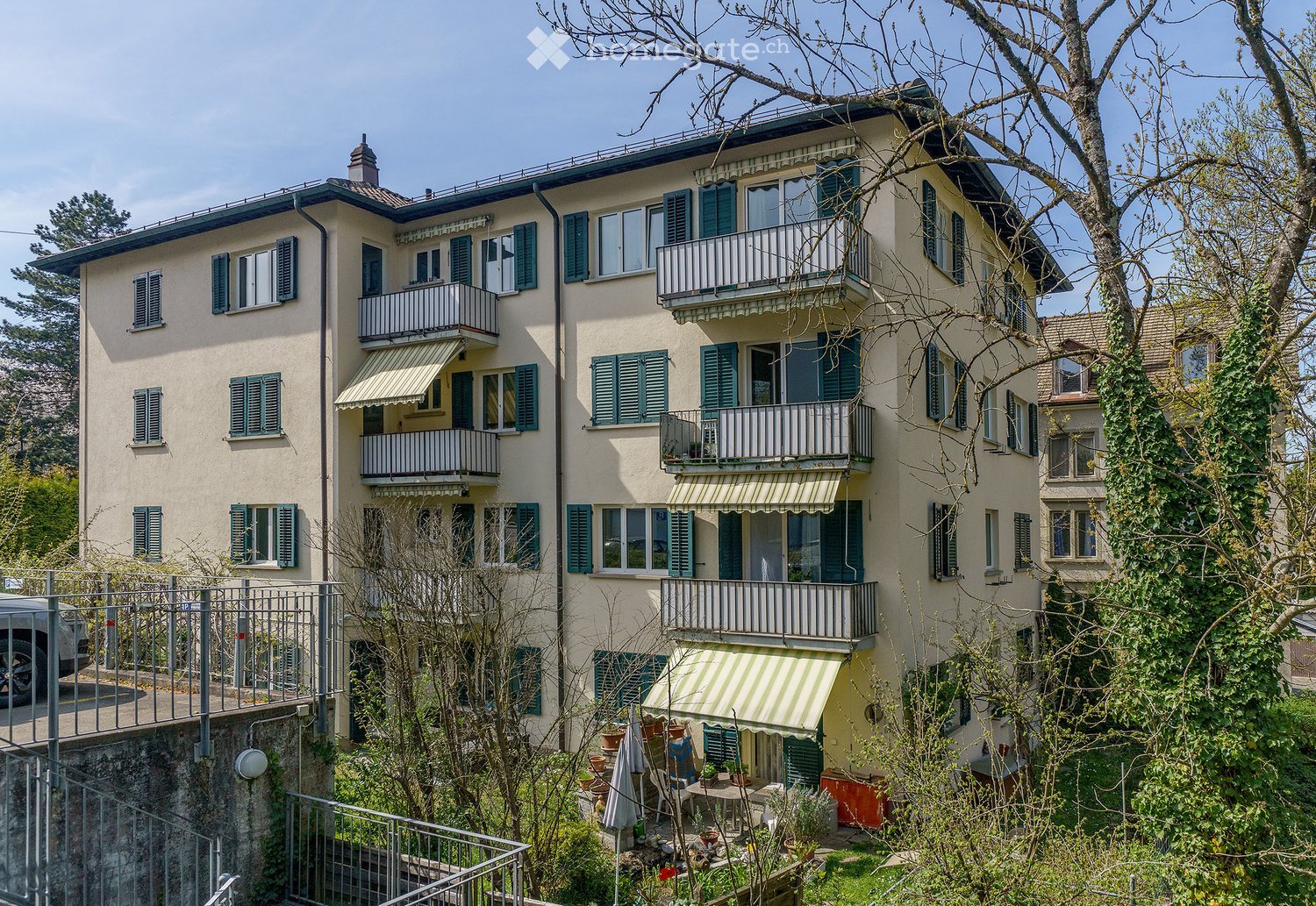 multi-storey building, beige exterior, green shutters, multiple balconies, small garden area, car parking area