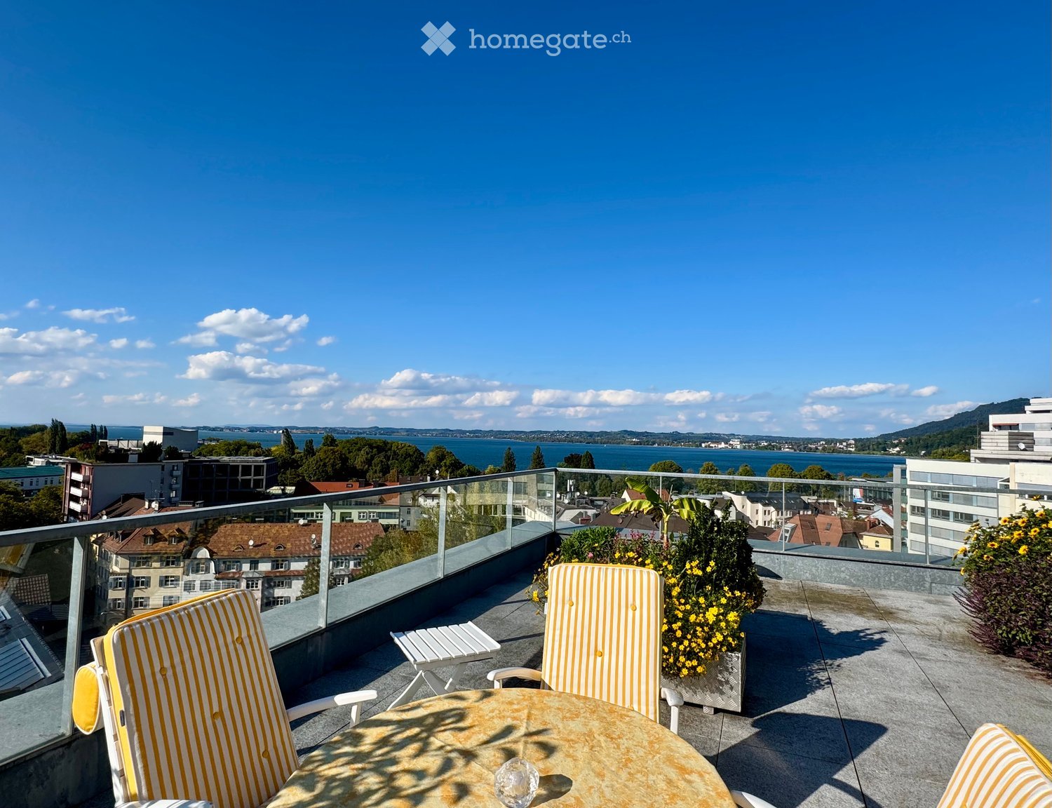 Terrace with table and chairs, yellow flowers, glass railing, view of the city and lake