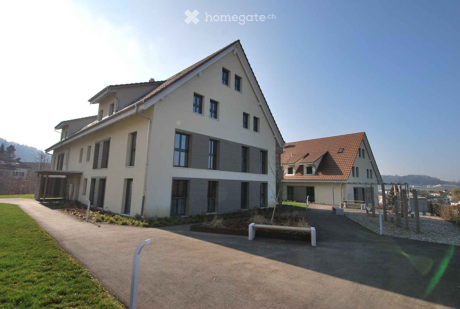 Two-story house, beige exterior, brown roof, multiple windows, driveway, small garden, bench