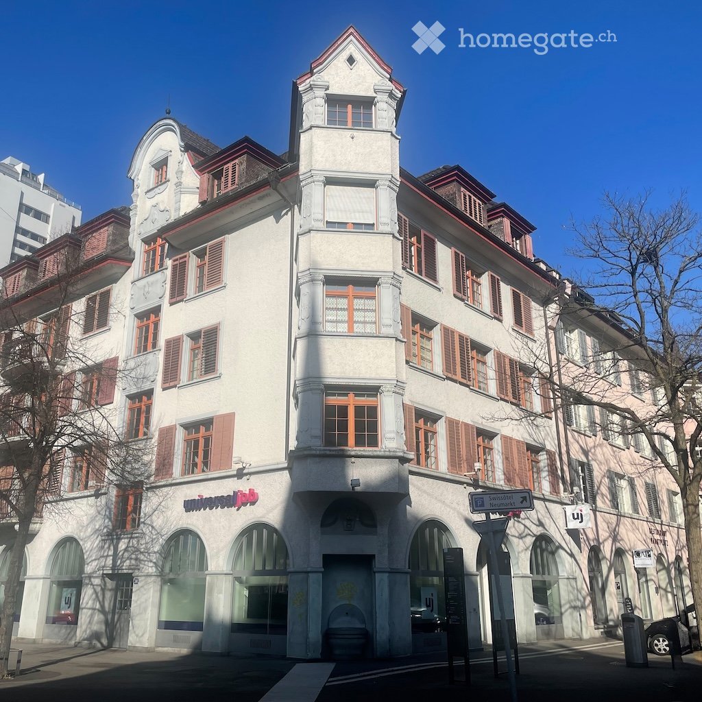 A multi-story building with white walls and red trim, arched windows, and a sign reading 'Universal Job'. The building has a modern architectural design with a combination of glass and metal elements. There are trees and a streetlight in front of the buil