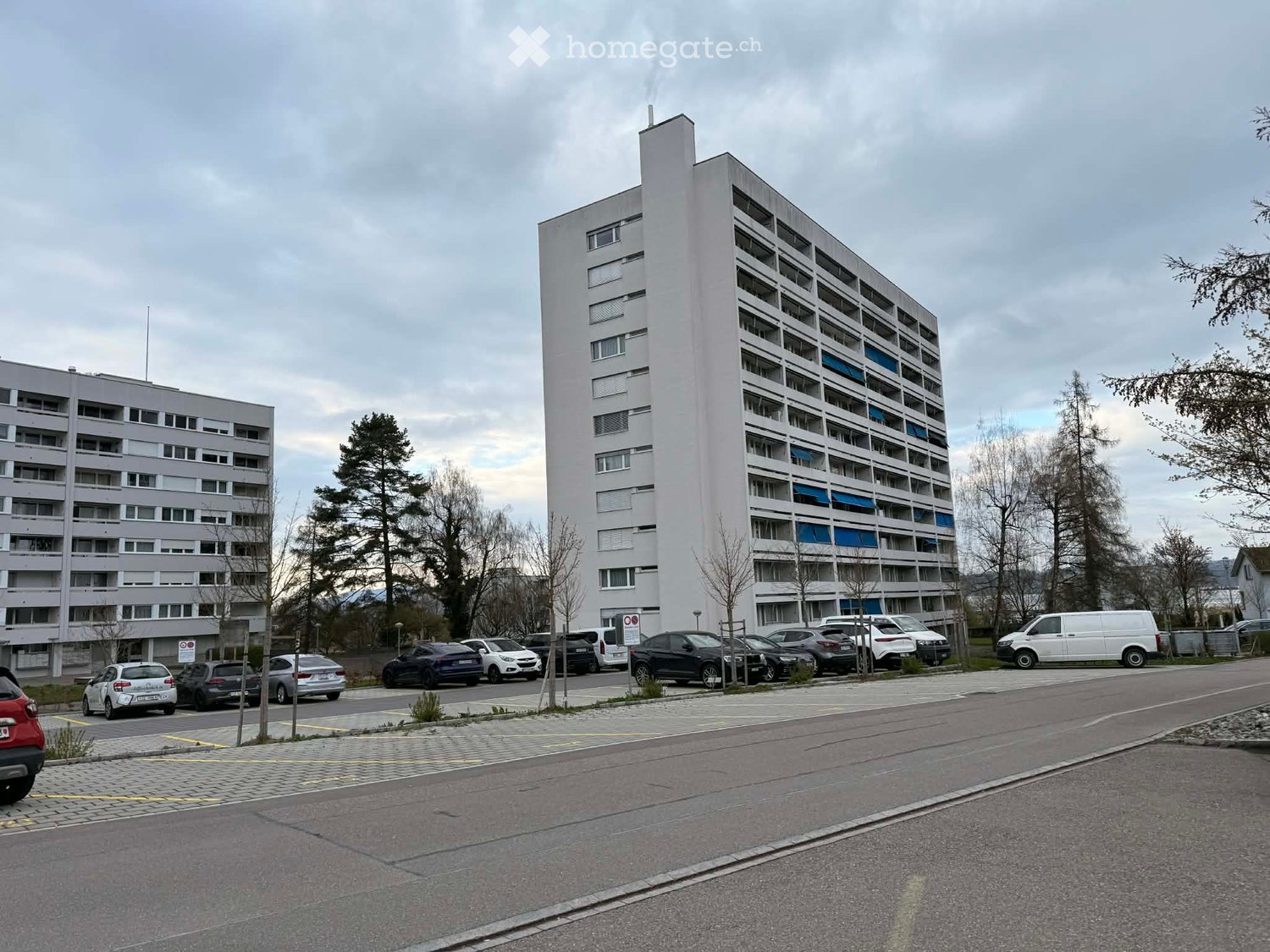 A tall white building with many windows and balconies, surrounded by trees and parked cars.