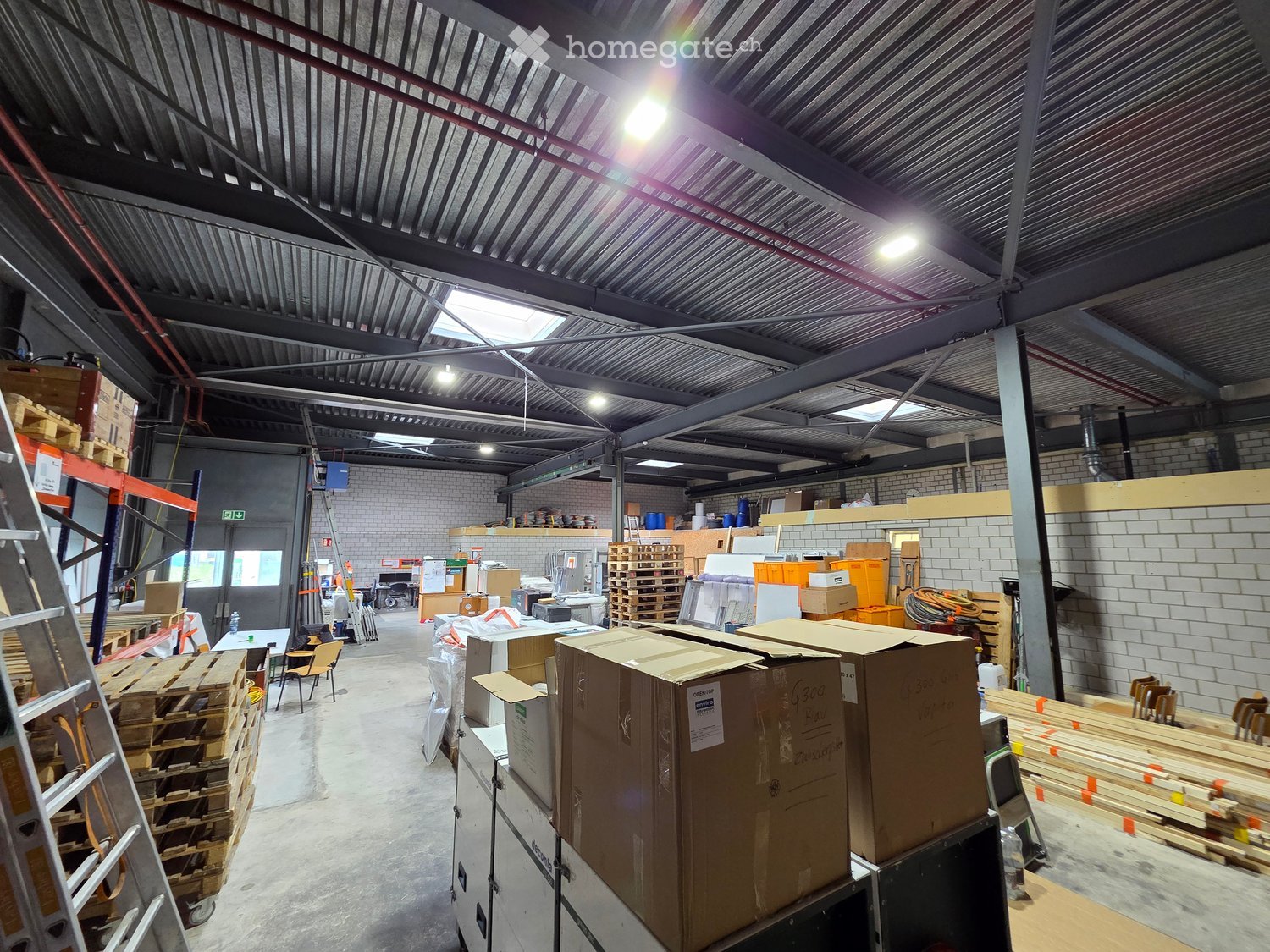 Warehouse with metal ceiling, brick walls, storage boxes, pallets, shelves, and various items