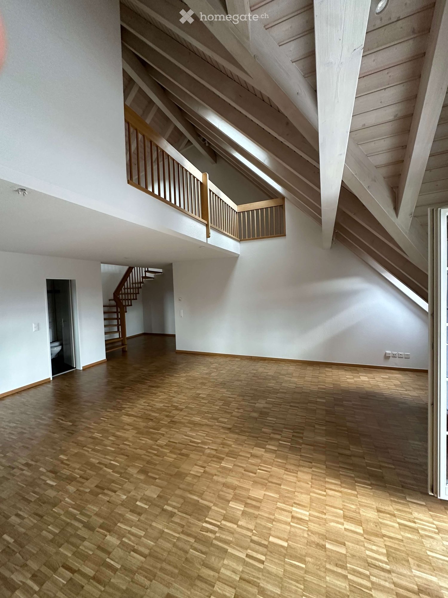 Empty room with wooden parquet flooring, white walls, wooden beams, and a wooden staircase leading to a balcony.