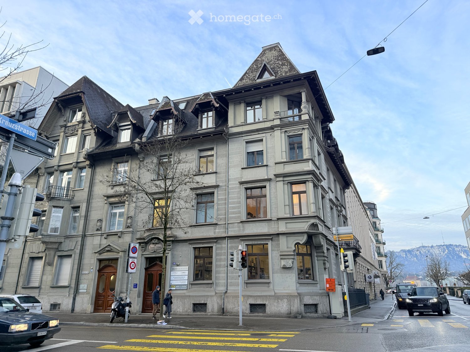 3 story building, gray exterior, multiple windows, two doors, street sign, cars, people walking