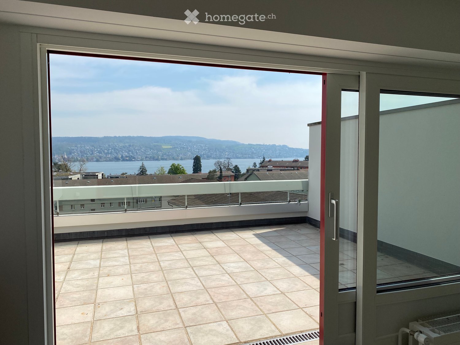 Open sliding door leading to a balcony with a tiled floor and a glass railing, overlooking a cityscape and a lake.
