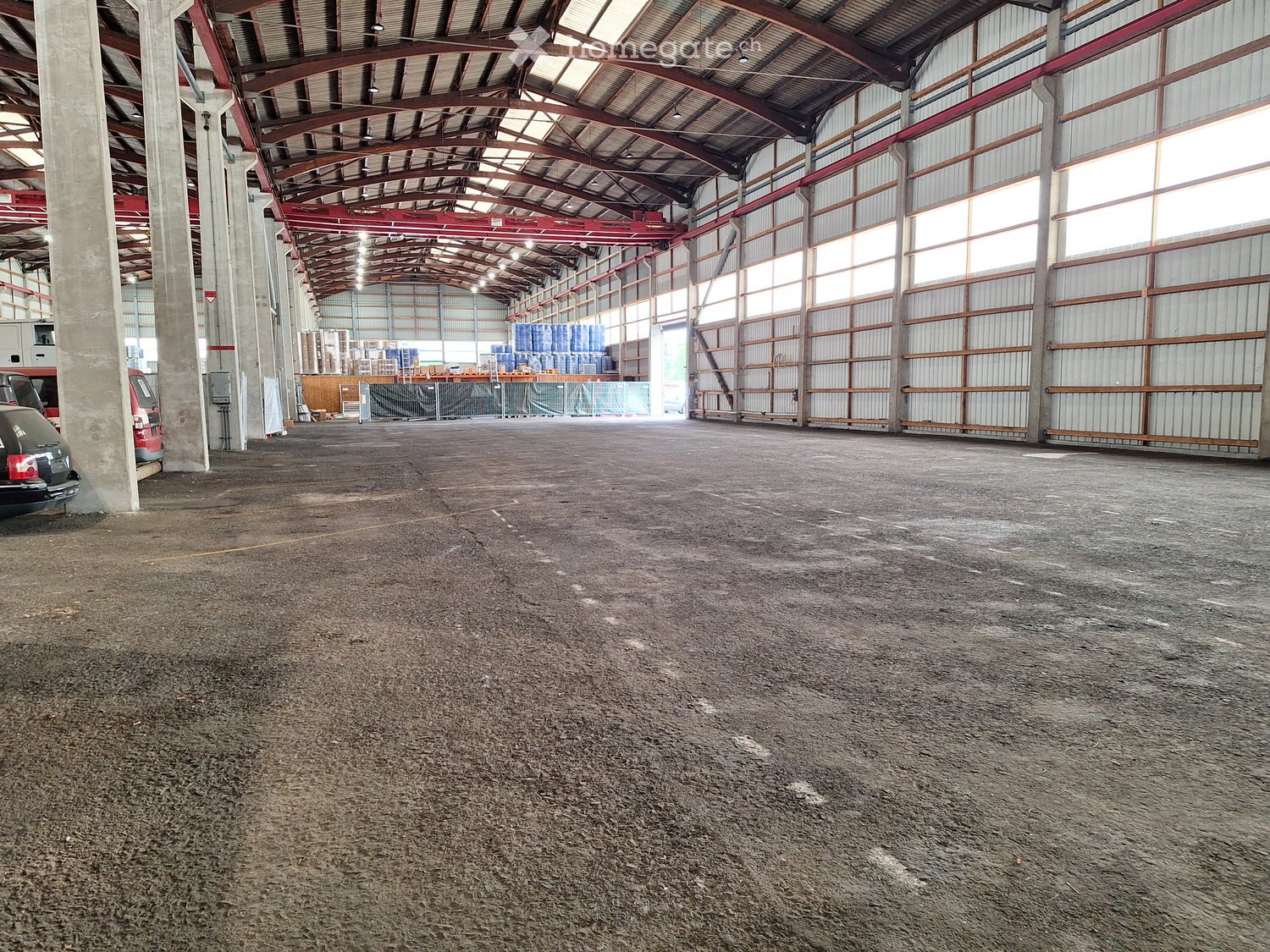 Large industrial warehouse with a high, metal-framed ceiling, concrete floor, and scattered storage items in the background.