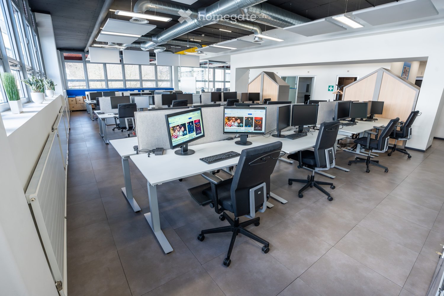 Open office space, desks, monitors, keyboards, chairs, windows, plants, air conditioning units