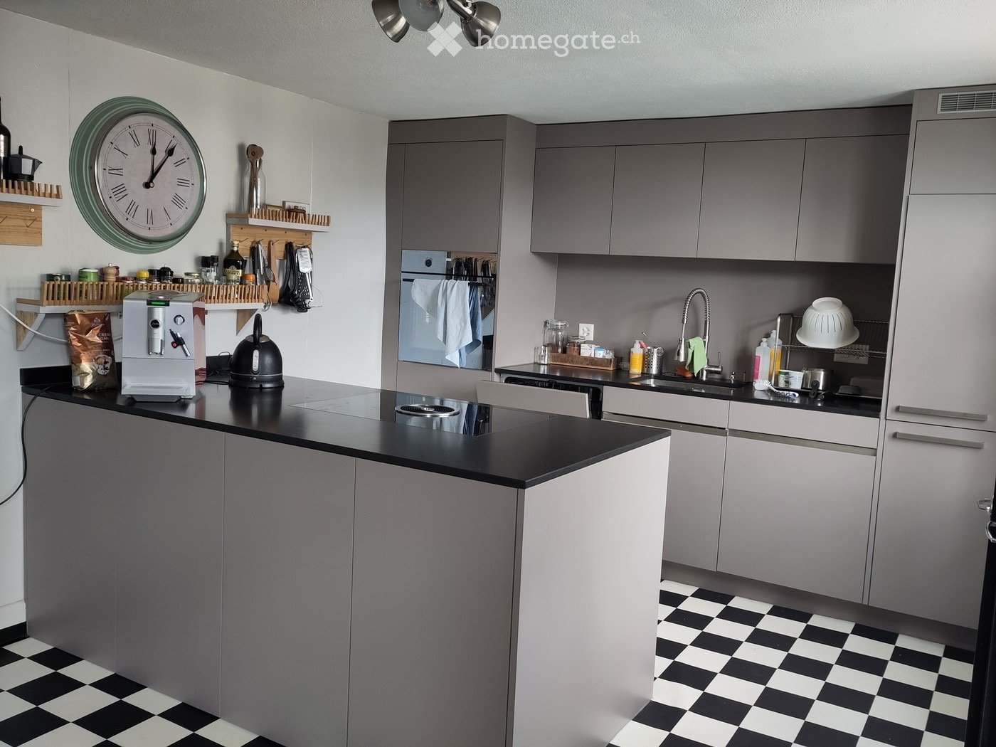 modern kitchen with island, black and white checkered floor, gray cabinets, black countertops, stainless steel appliances, large clock on wall
