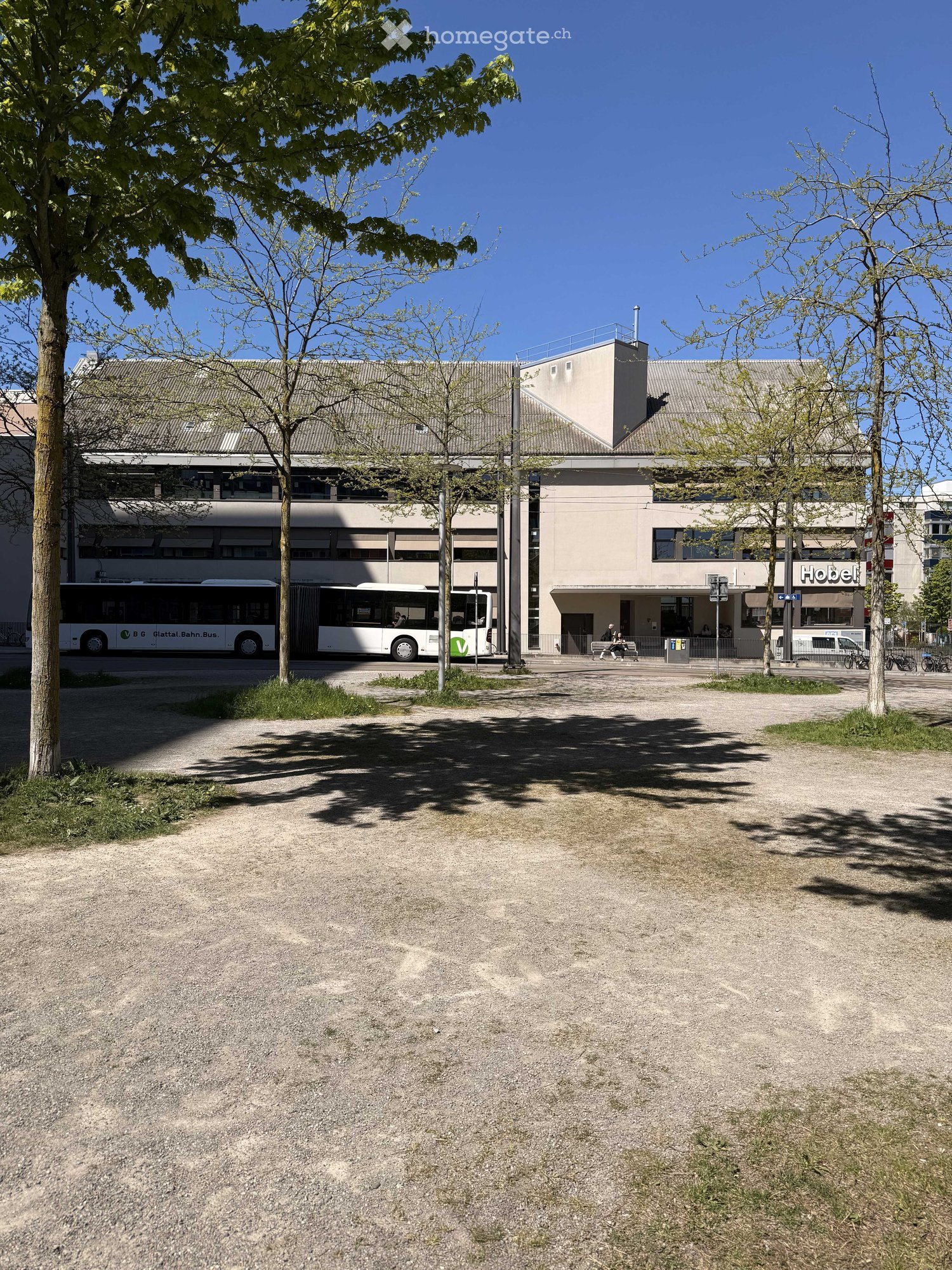 A large building with a bus stop in front, trees, and a parking lot.
