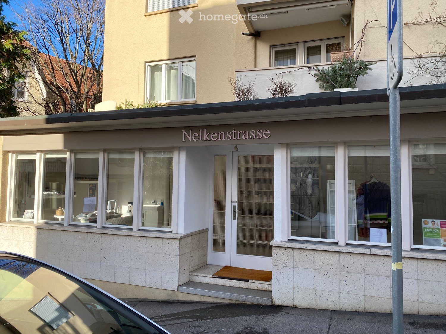 A commercial building with a sign reading 'Nelkenstrasse' above the entrance. The building has a modern design with glass windows and a glass door. There are potted plants on the balcony and a car parked in front.