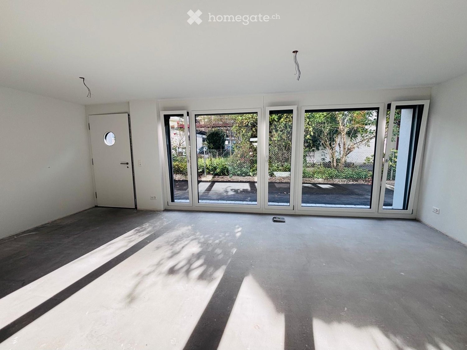 Empty room with white walls, wooden flooring, large sliding glass doors leading to an outdoor area, and a single door on the left side.