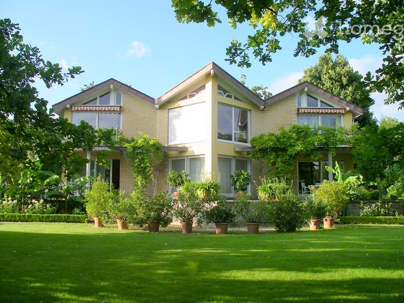 Two-story house with a yellow exterior, large windows, and a well-maintained garden with various potted plants.