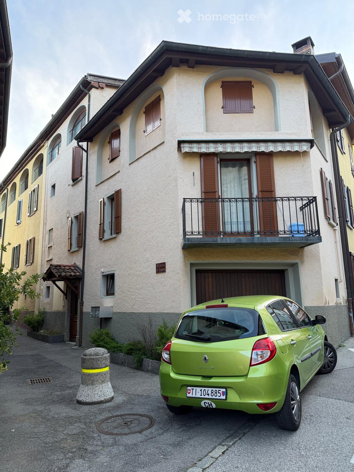 A three-story building with beige walls and brown shutters. A green car is parked in front of the building.