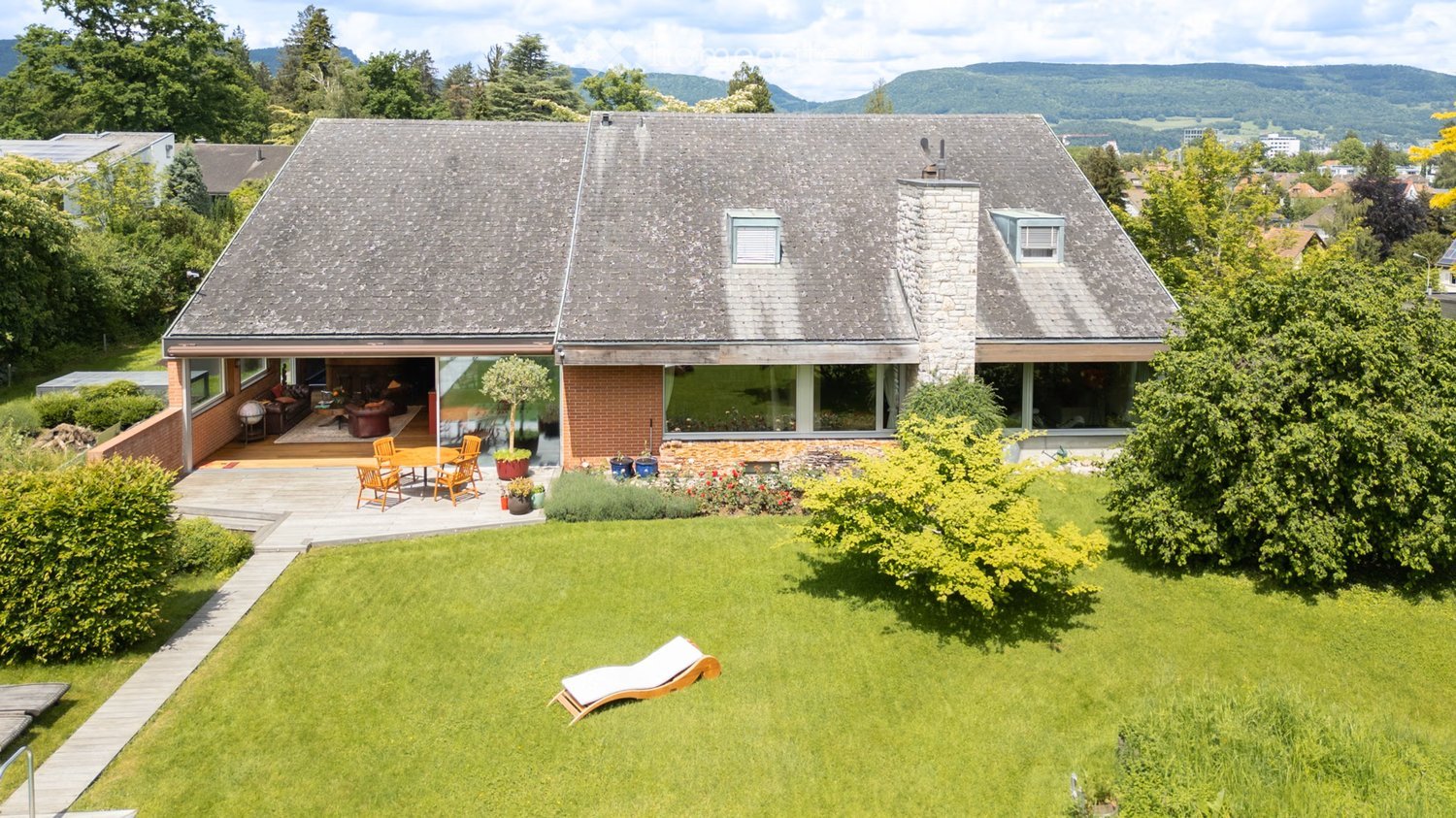 Detached house with gray roof, brick walls, glass windows, wooden deck, garden with lounger, outdoor table and chairs