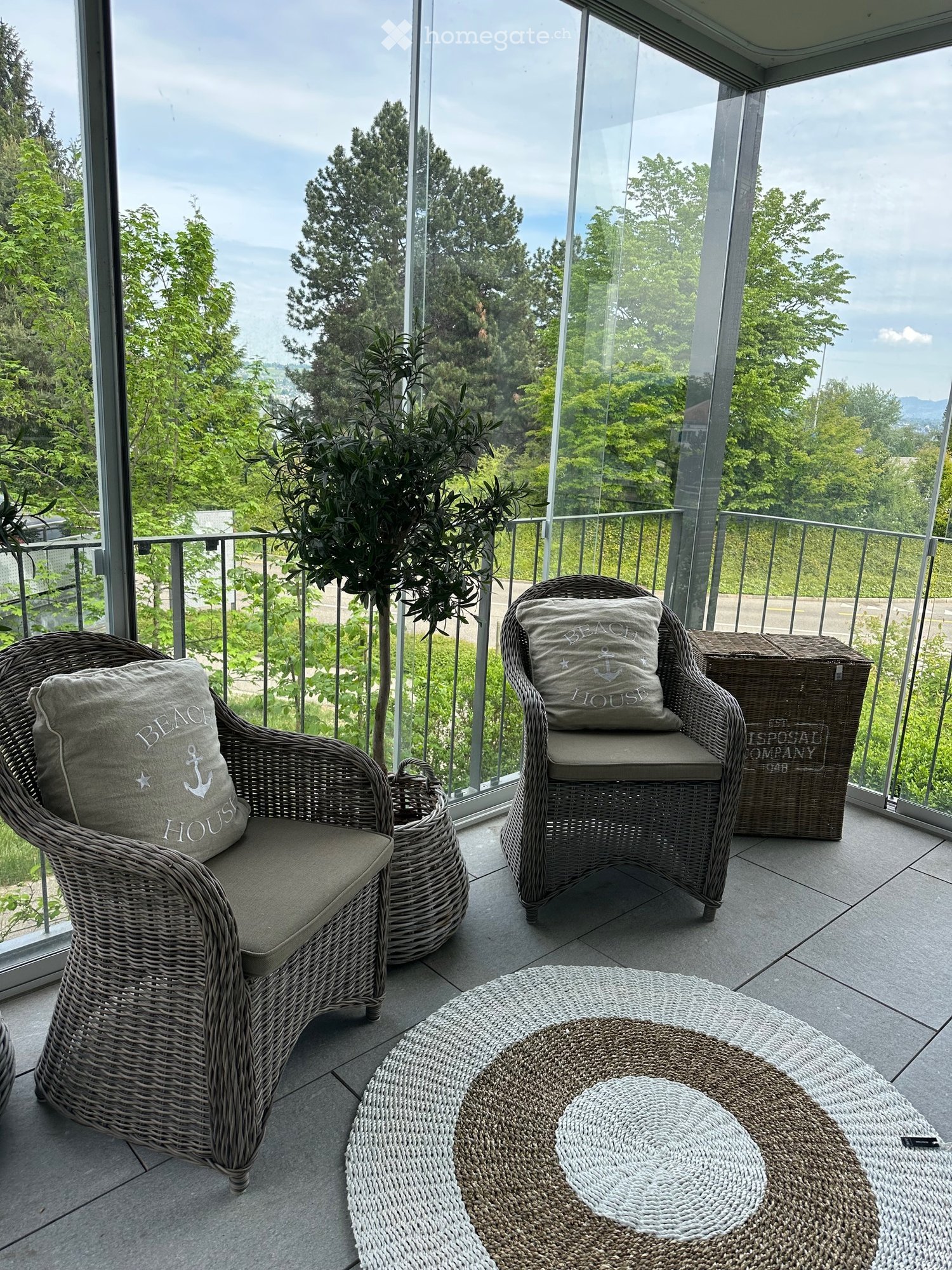 Balcony / Terrace. A balcony with a view of trees and a road, featuring two wicker chairs with cushions, a potted plant, a storage box, and a round rug.