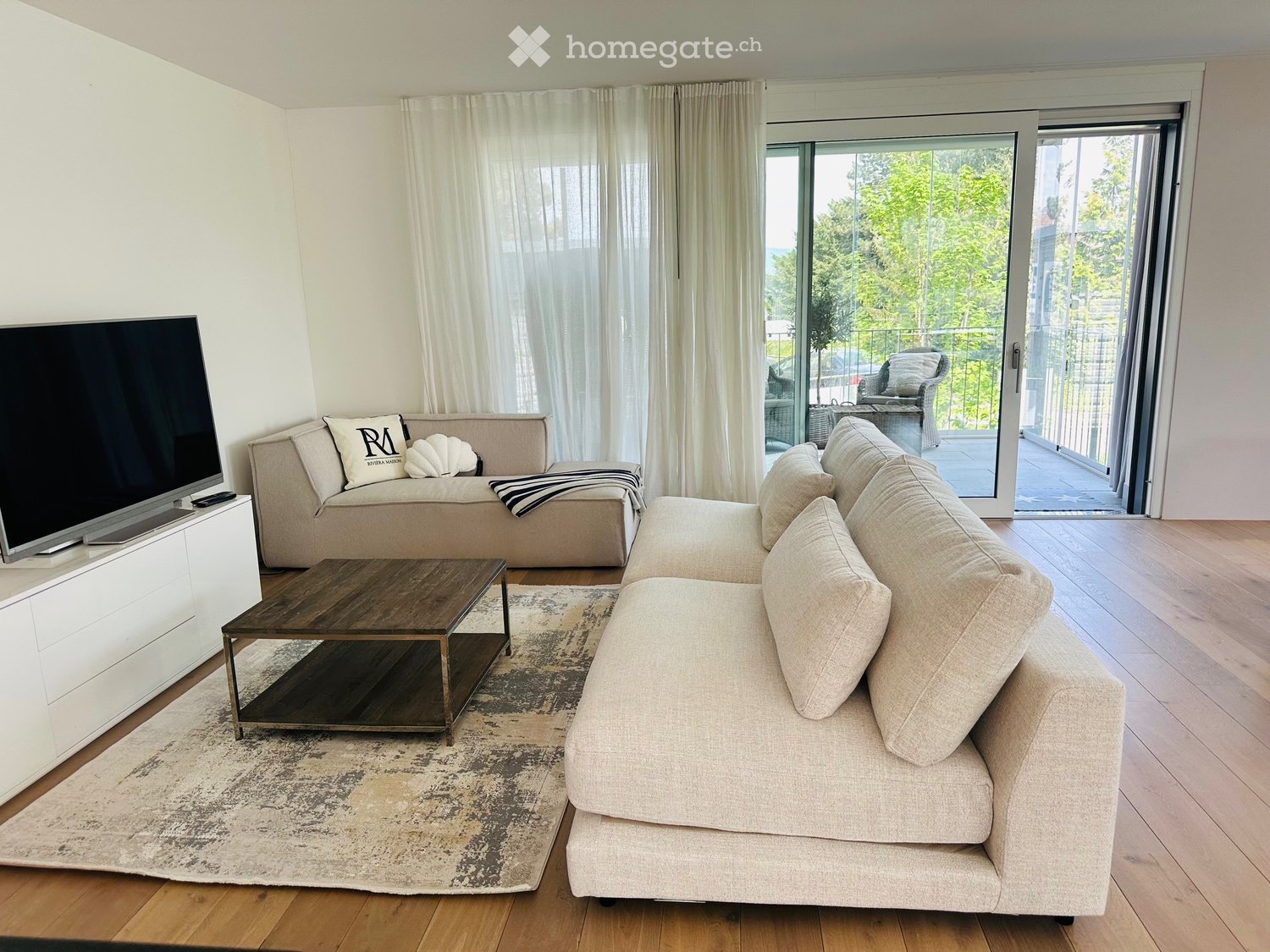 Living Room. Modern living room with a large L-shaped sofa, a wooden coffee table, and a flat-screen TV on an entertainment center. A decorative pillow and a throw pillow are on the sofa. A rug covers part of the wooden floor. The room has large glass doo