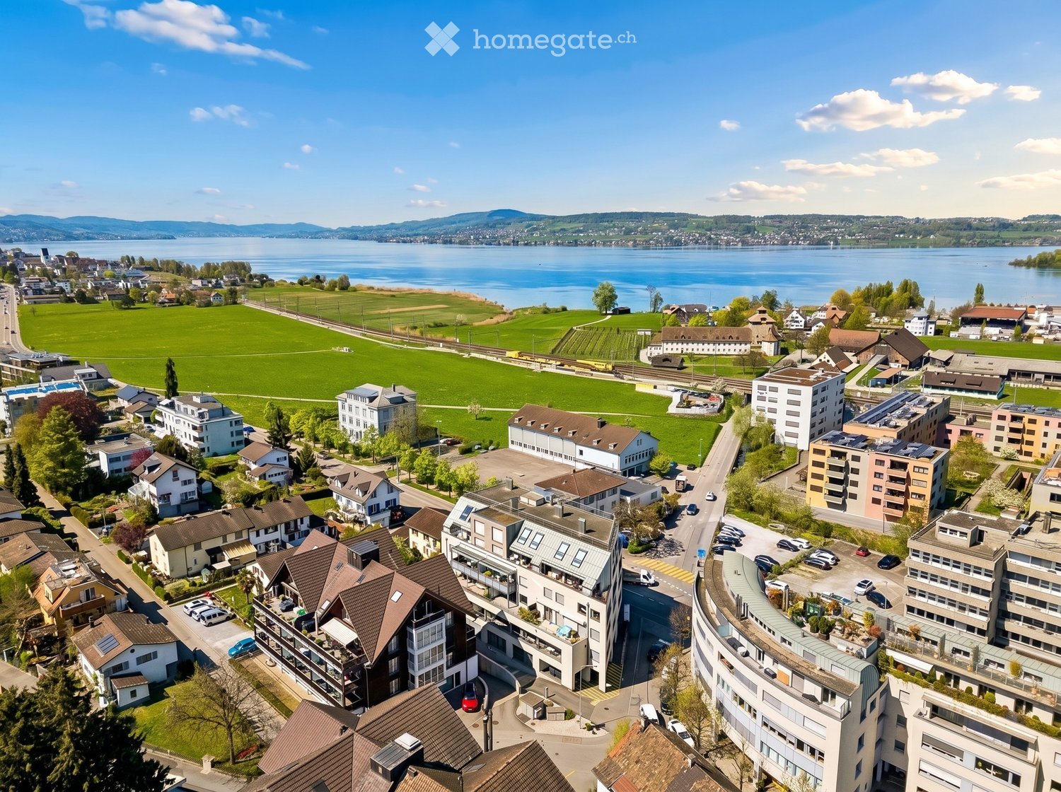 Building. Aerial view of a residential area with various buildings, a large green field, and a body of water in the background.