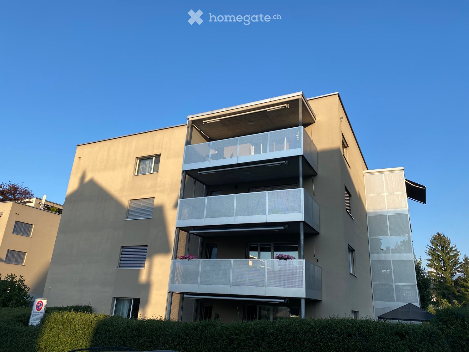 A modern apartment building with three floors, each floor having multiple balconies, each with railings, there is a shaded area and greenery in the front.