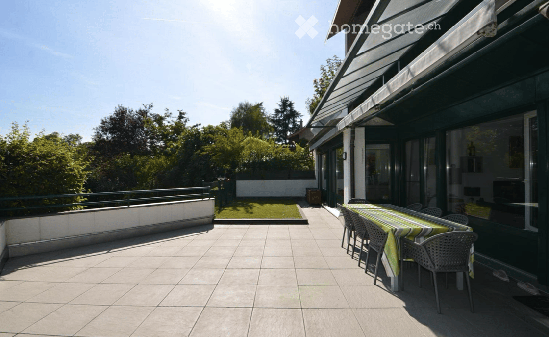 Outdoor terrace with a table and chairs, covered by an umbrella, surrounded by a garden.