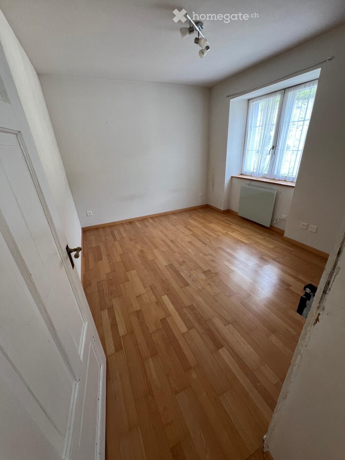 Empty room with wooden flooring, white walls, and a window with white curtains. A radiator is visible under the window, and a door is on the left side.