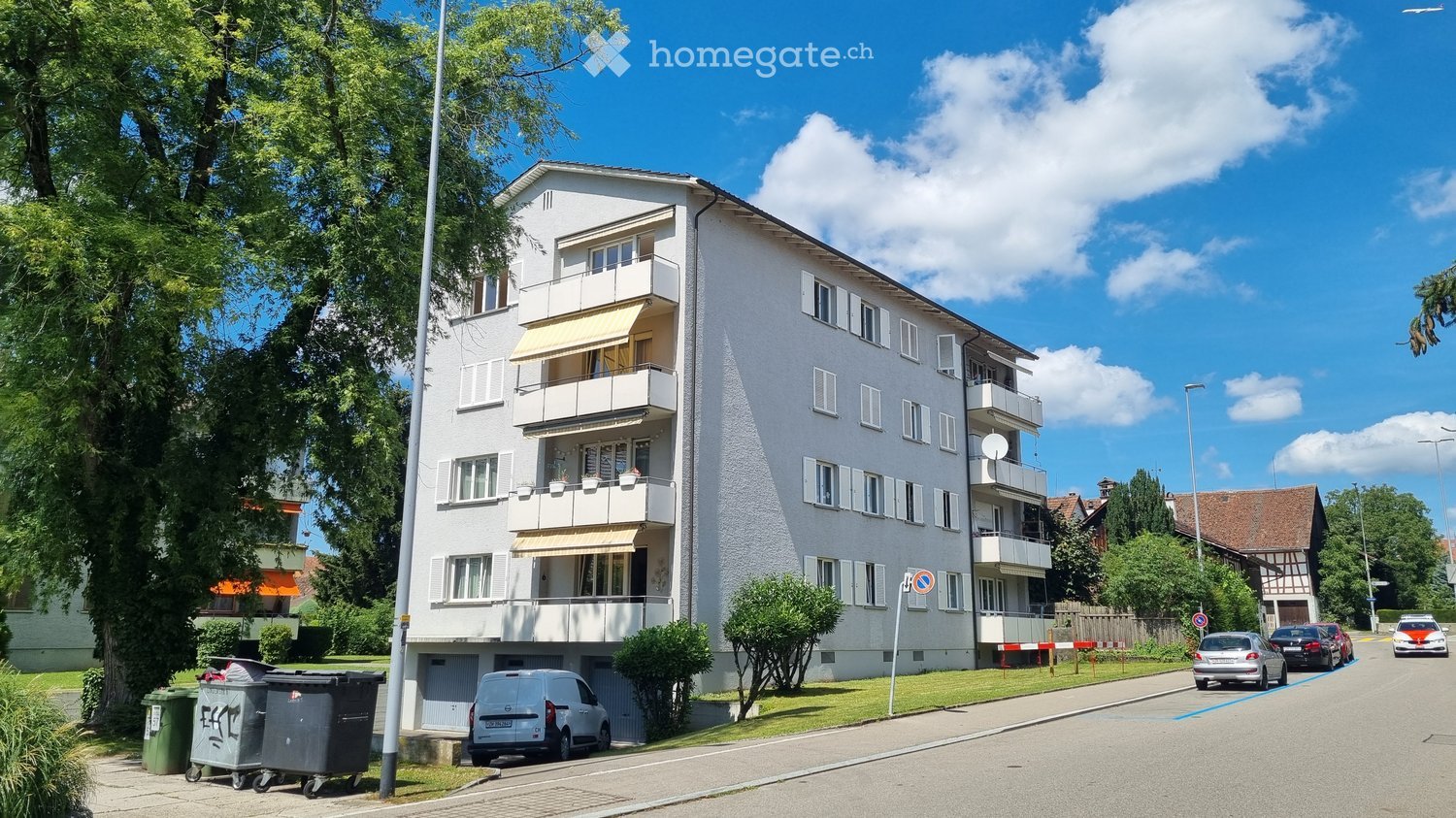 A multi-story residential building with balconies and awnings, situated in a suburban area with trees and parked cars.