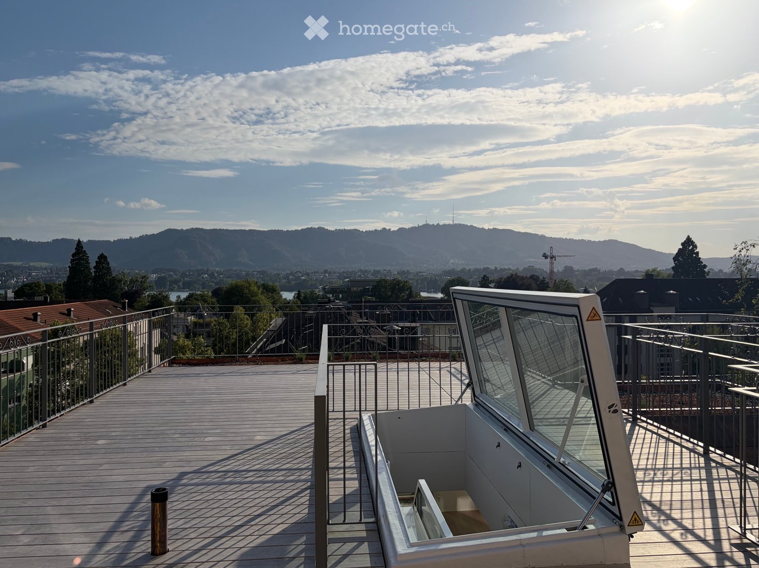 Wooden deck with metal railing, glass-enclosed elevator shaft, scenic city and mountain view.