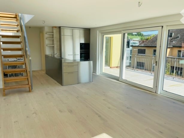 Empty room with wooden floor, white walls, and sliding glass doors leading to a balcony. A staircase is on the left side, and a kitchen area is on the right side.