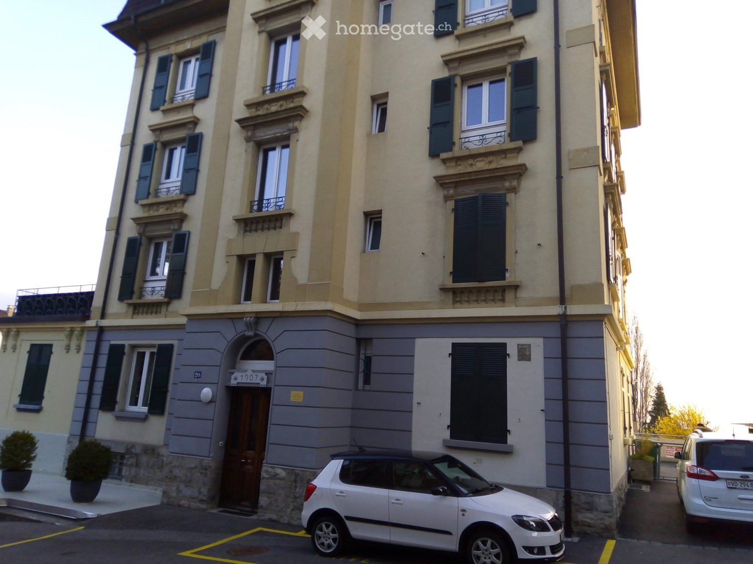 Apartment building with multiple windows and doors, car parked in front, potted plants at the entrance