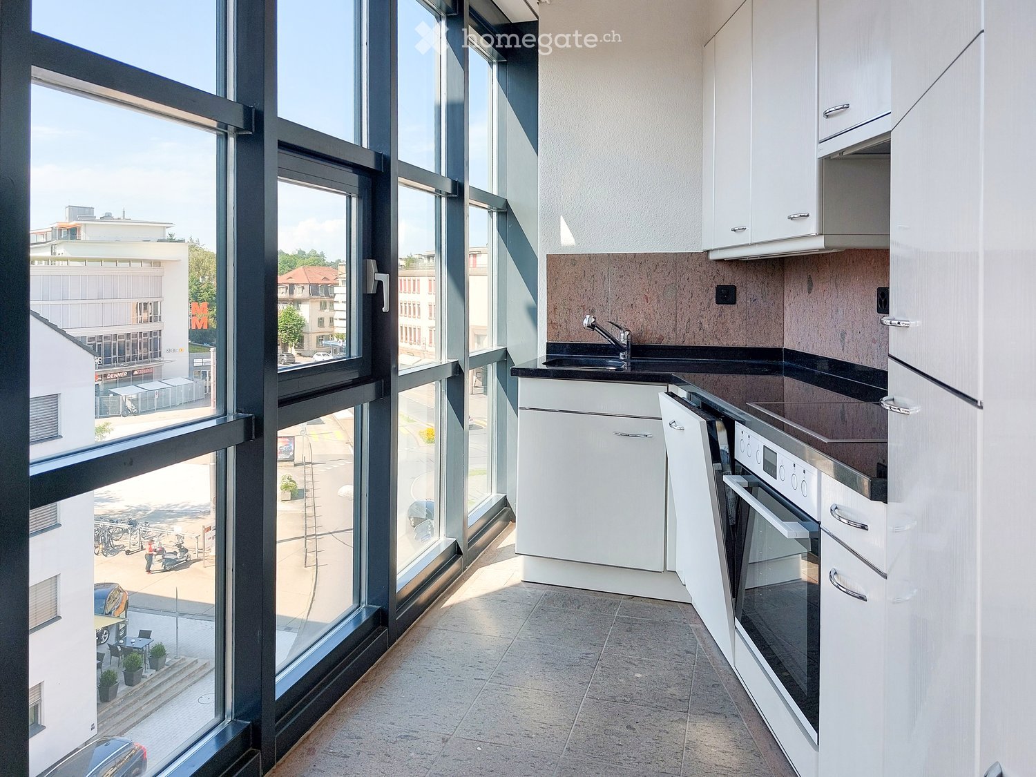 modern kitchen with large windows, white cabinets, black countertops, stainless steel sink, oven, refrigerator, and a view of the city