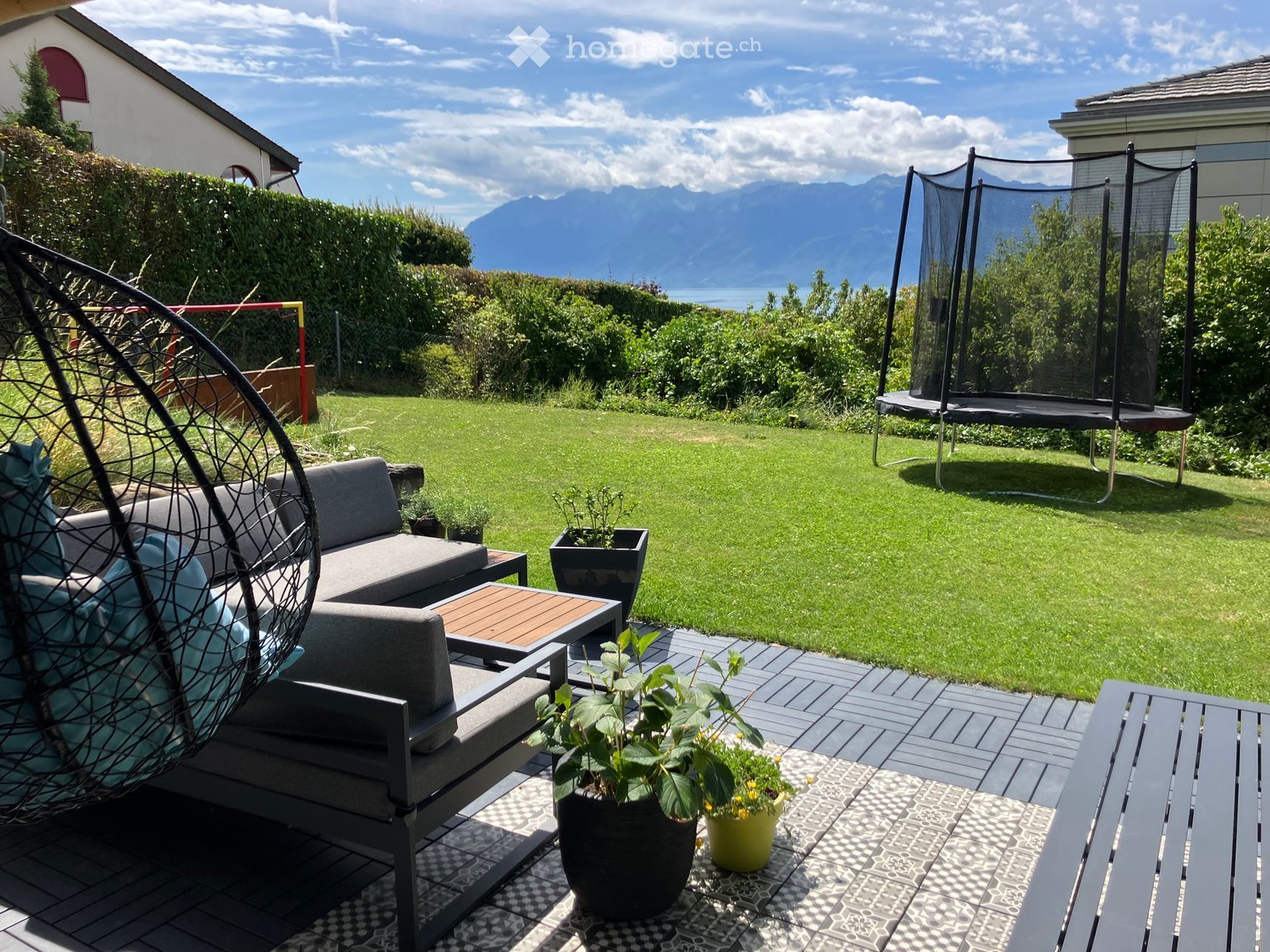 Outdoor seating area with a couch, table, and a hammock. Potted plants and a trampoline are in the garden. The garden has a view of the mountains.