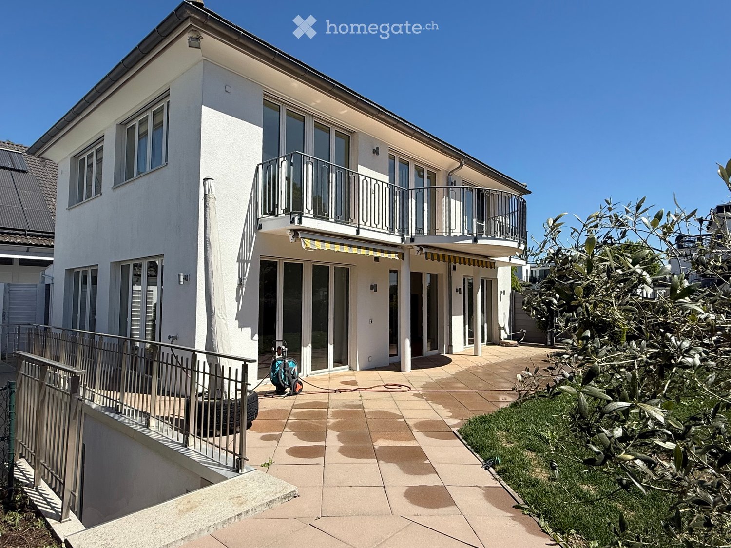Modern white house with two floors, large windows, and a balcony with metal railings. The entrance has a paved walkway leading to the front door.