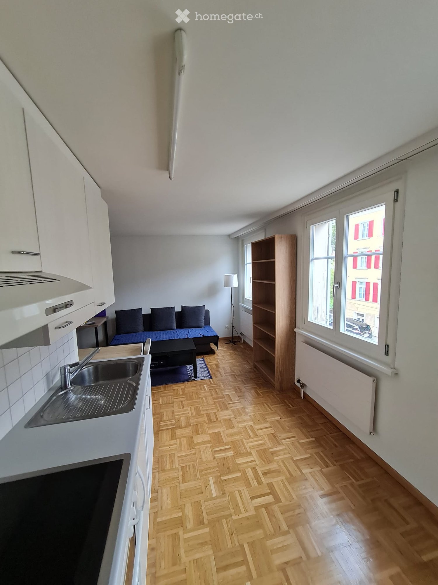 A modern, open-plan living and kitchen area with wooden herringbone flooring, white cabinets, a sofa, a small table, a sink, and a floor lamp.