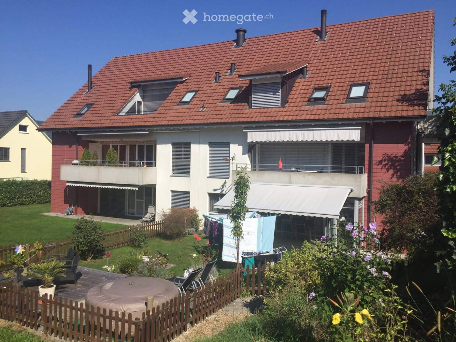 House. Two-story house with a red roof, white windows, and a balcony with awnings. The house is surrounded by a garden with a wooden fence, plants, and flowers. There is a patio with a round table and chairs, and a potted plant. A clothesline with clothes