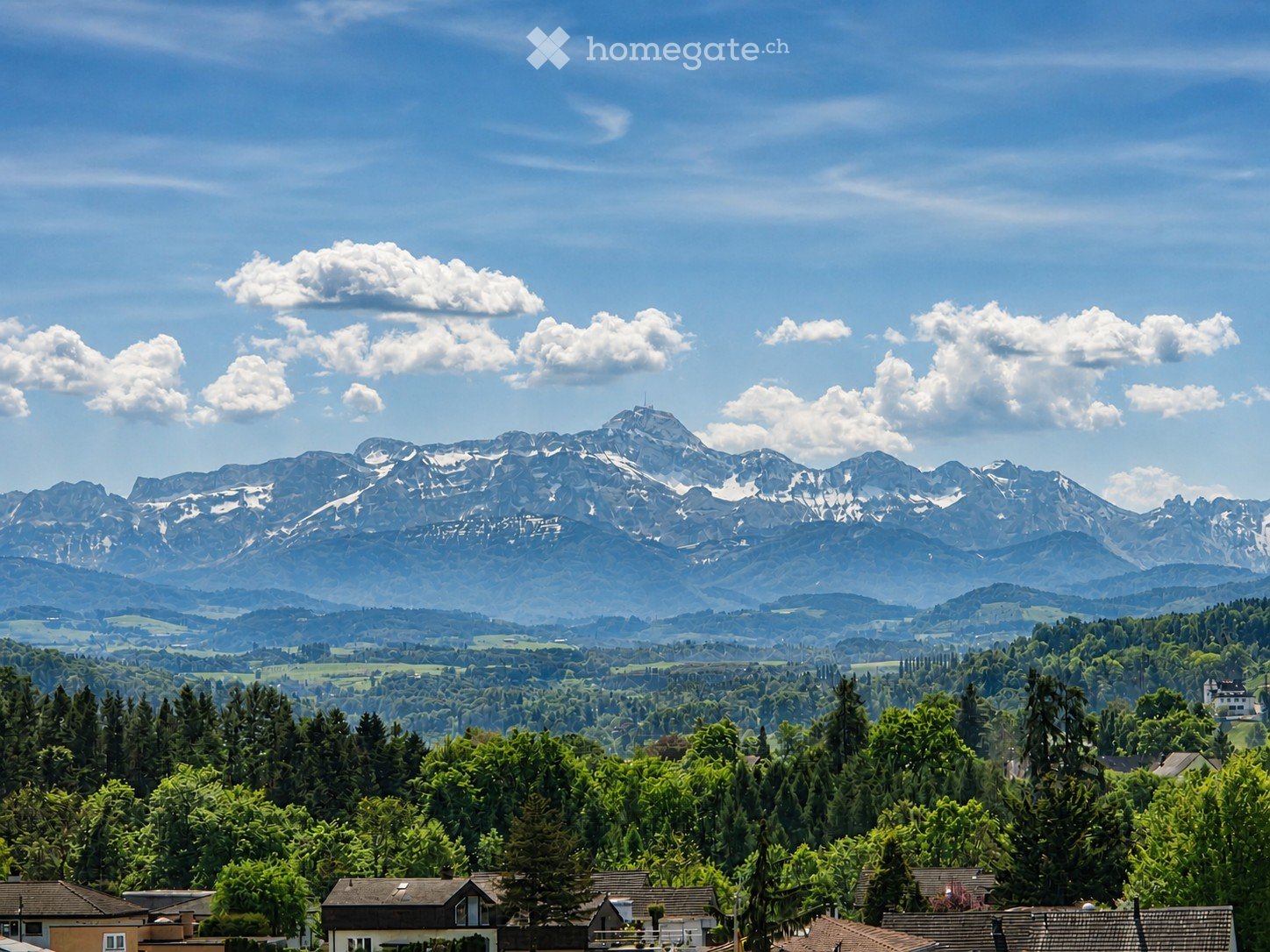Aerial view of a valley with a mountain range in the background, surrounded by lush green trees and houses.