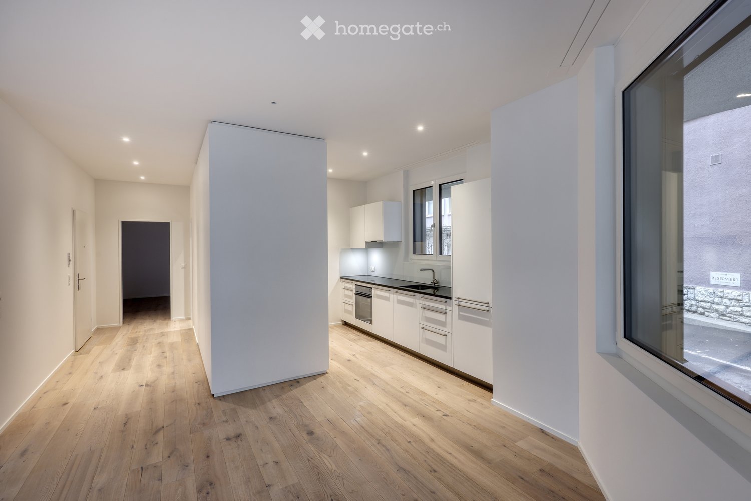An empty room with white walls, wooden floor, and a large window. The room has a modern design with a minimalist aesthetic.