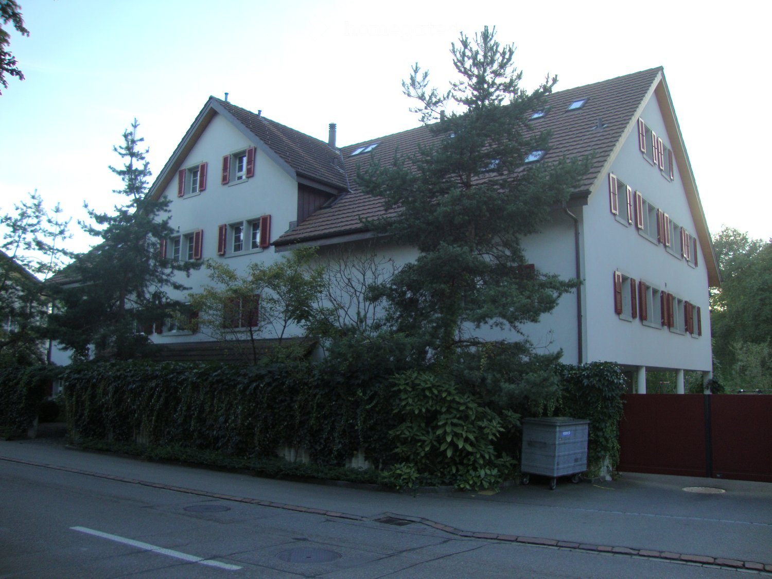 2 story house, white exterior, brown roof, multiple windows, red shutters, surrounded by trees and bushes, red gate