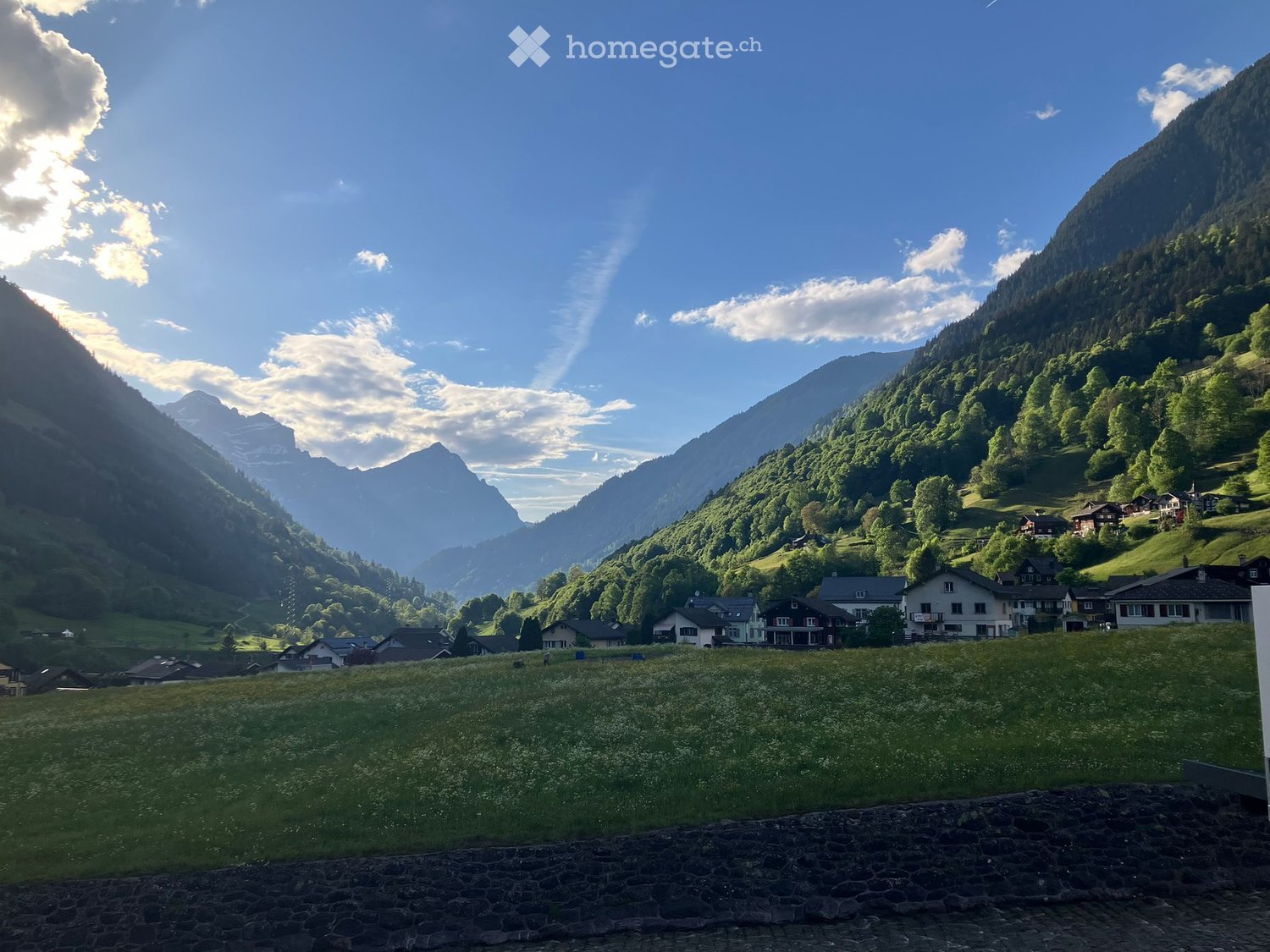 A picturesque alpine valley with scattered houses, lush green meadows, and towering mountains under a clear blue sky.