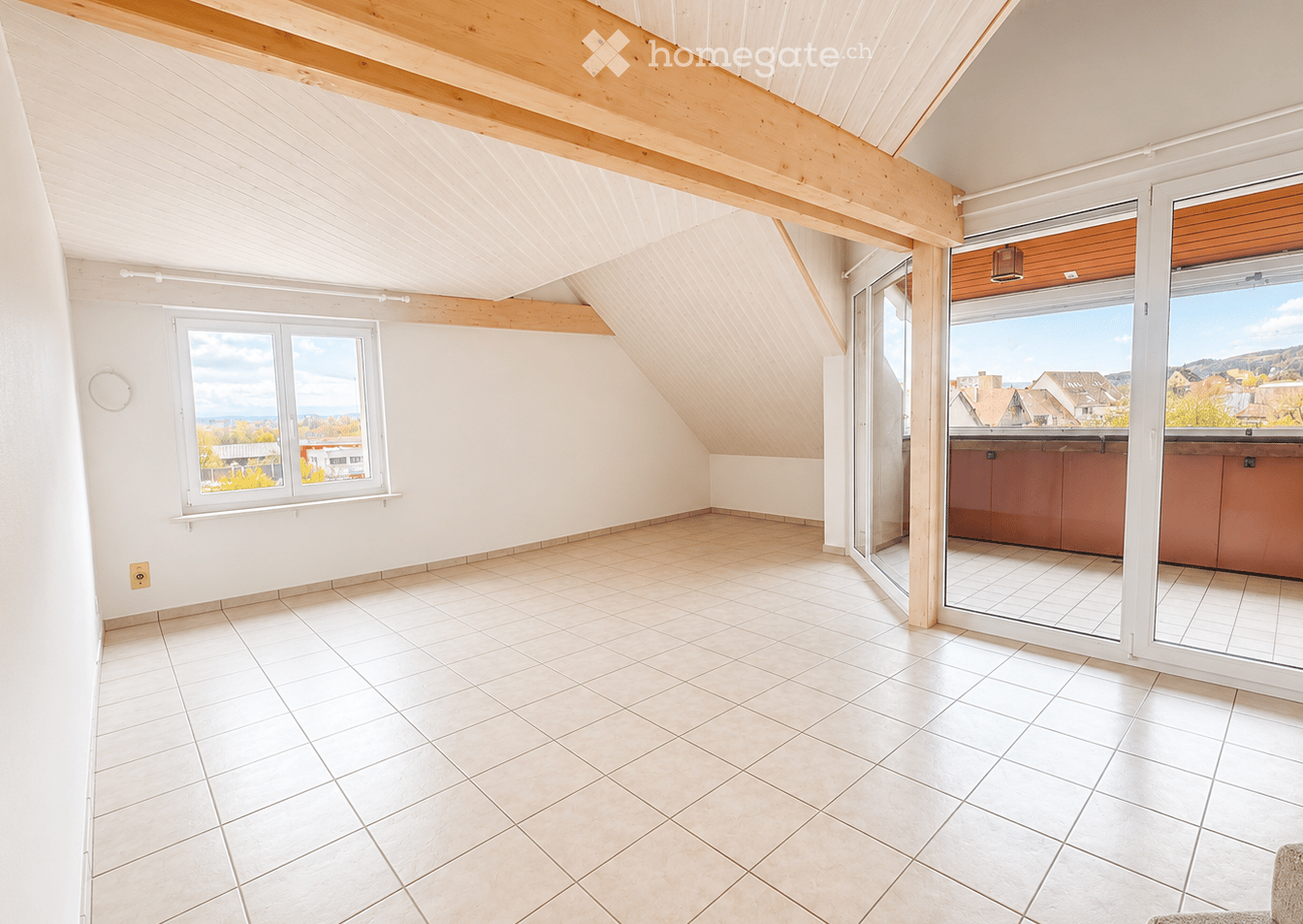 Bright, empty attic room with white walls, tiled floor, large windows, and a sliding glass door leading to a balcony with a scenic view.