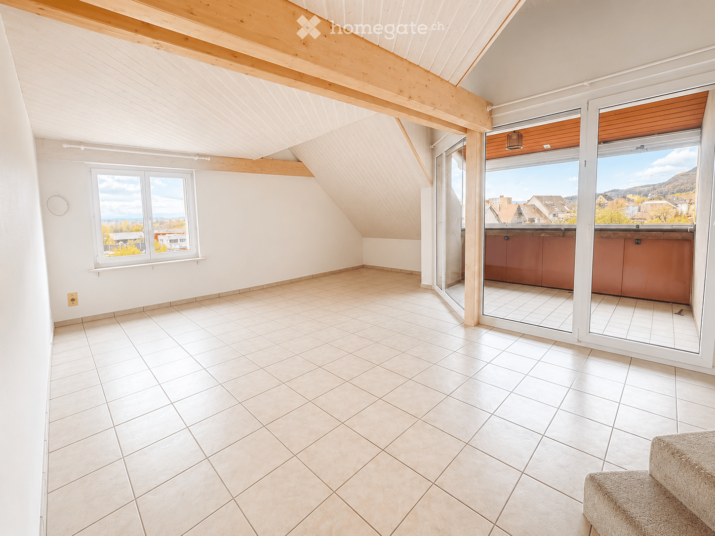 Bright, unfurnished attic room with wooden slanted ceilings, large windows, and a sliding glass door leading to a balcony with scenic views.