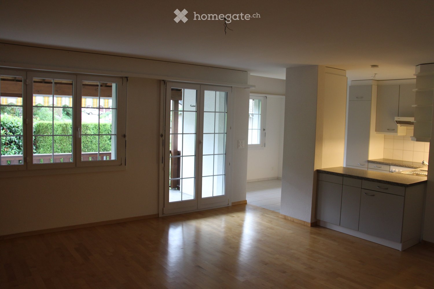 Unfurnished Room. Empty room with wooden floors, large windows, and sliding glass doors leading to a balcony. The room has white walls and a modern kitchen area with cabinets and countertops.