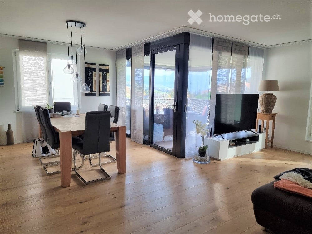 Open-plan living and dining area with wooden flooring, large glass sliding doors leading to a balcony, a dining table with chairs, a television on a stand, a lamp, and a sofa.